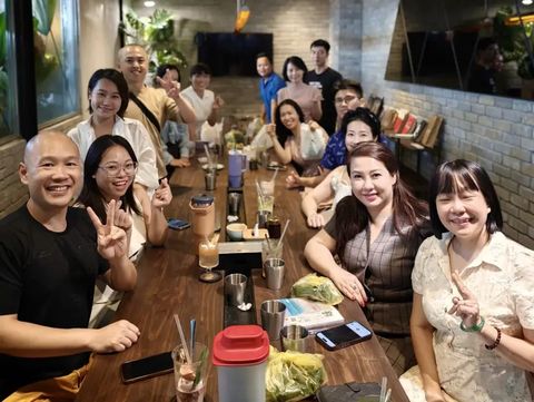 Một nhóm người ngồi quanh chiếc bàn dài trong quán cà phê, cười và giơ tay chào về phía máy ảnh.