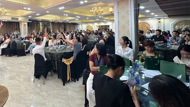 Hội trường đông người ngồi quanh các bàn tròn, nhiều người giơ tay.