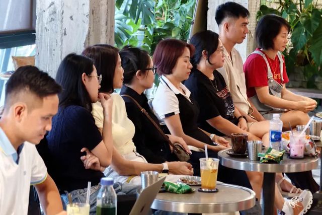 Một hàng người ngồi bên những bàn cà phê nhỏ, chăm chú lắng nghe; phía trước có một máy tính xách tay.