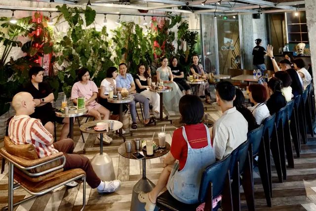 Khoảng hai mươi người ngồi thành vòng quanh những bàn nhỏ trong một quán cà phê nhiều cây xanh; một người giơ tay.